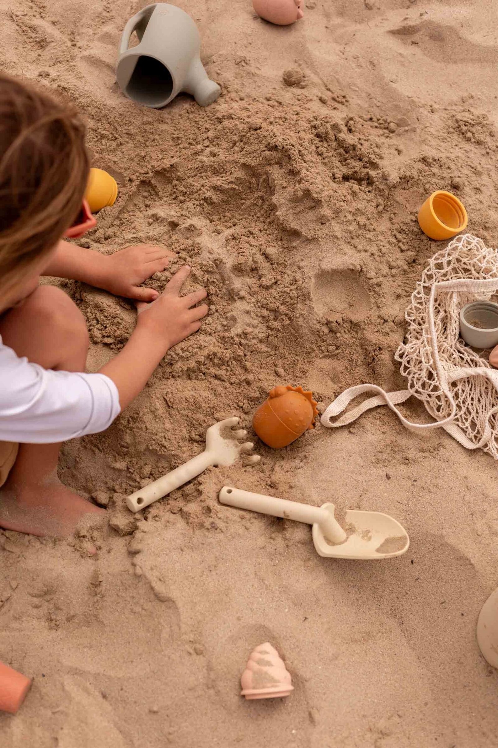 child playing at the beach in the sand with his bands and sand toys surrounding him